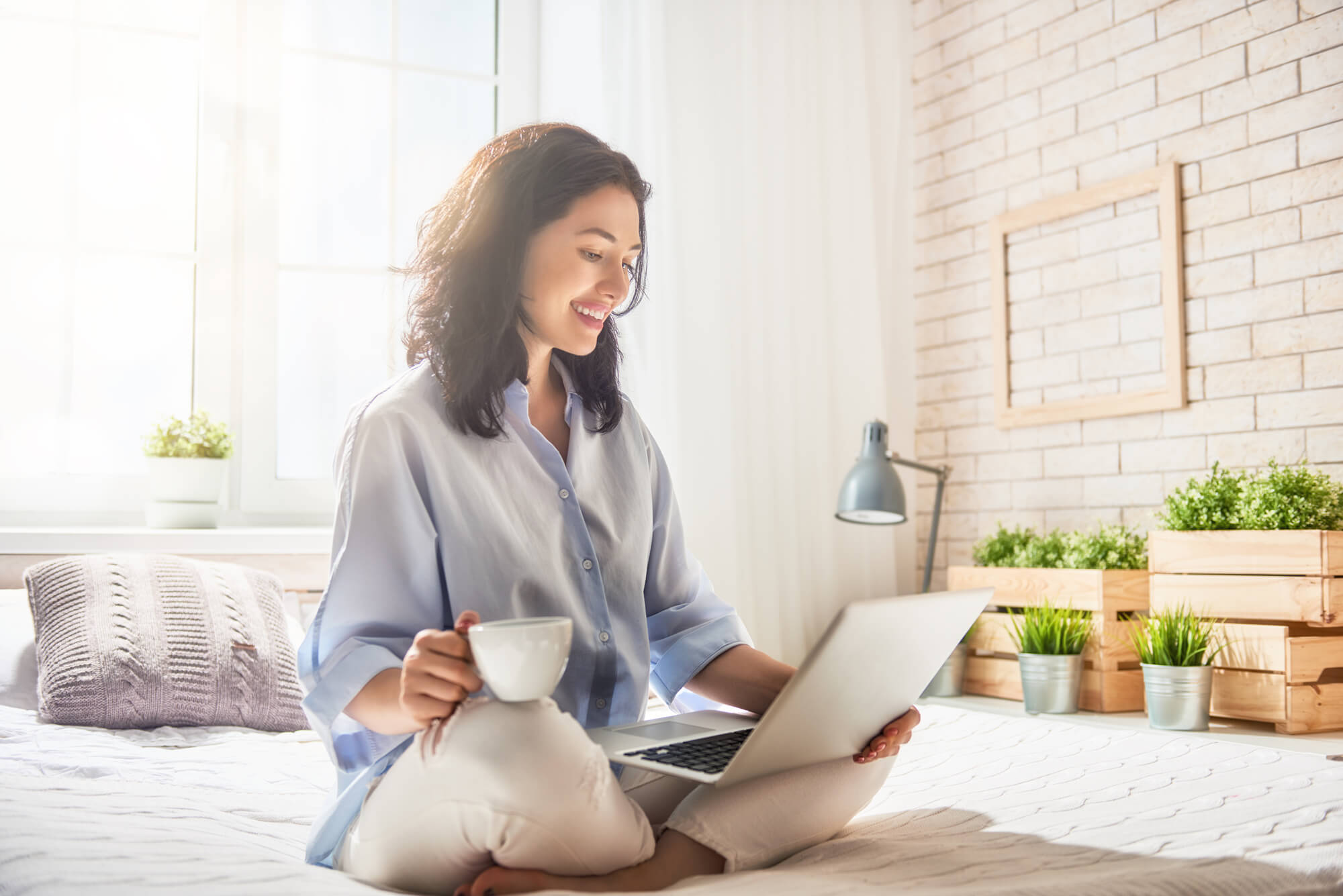 Une femme consulte son portable assis en indien sur son lit avec un café à la main